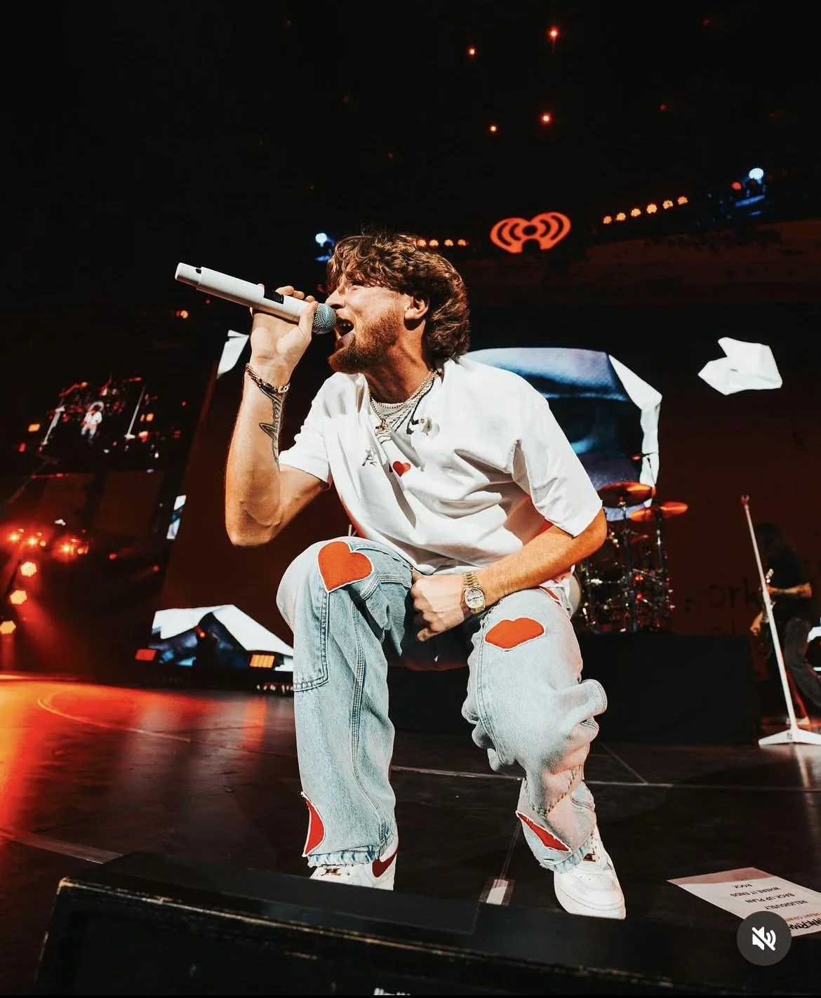 A male singer performs on stage, kneeling with one knee on the ground, holding a microphone close to his mouth. He is wearing a white t-shirt with red heart patches, distressed jeans, and white sneakers with red accents. The stage is lit with red and orange lights, and there is a drum set and other band members in the background. The singer has brown hair, a beard, and is wearing a watch and jewelry.