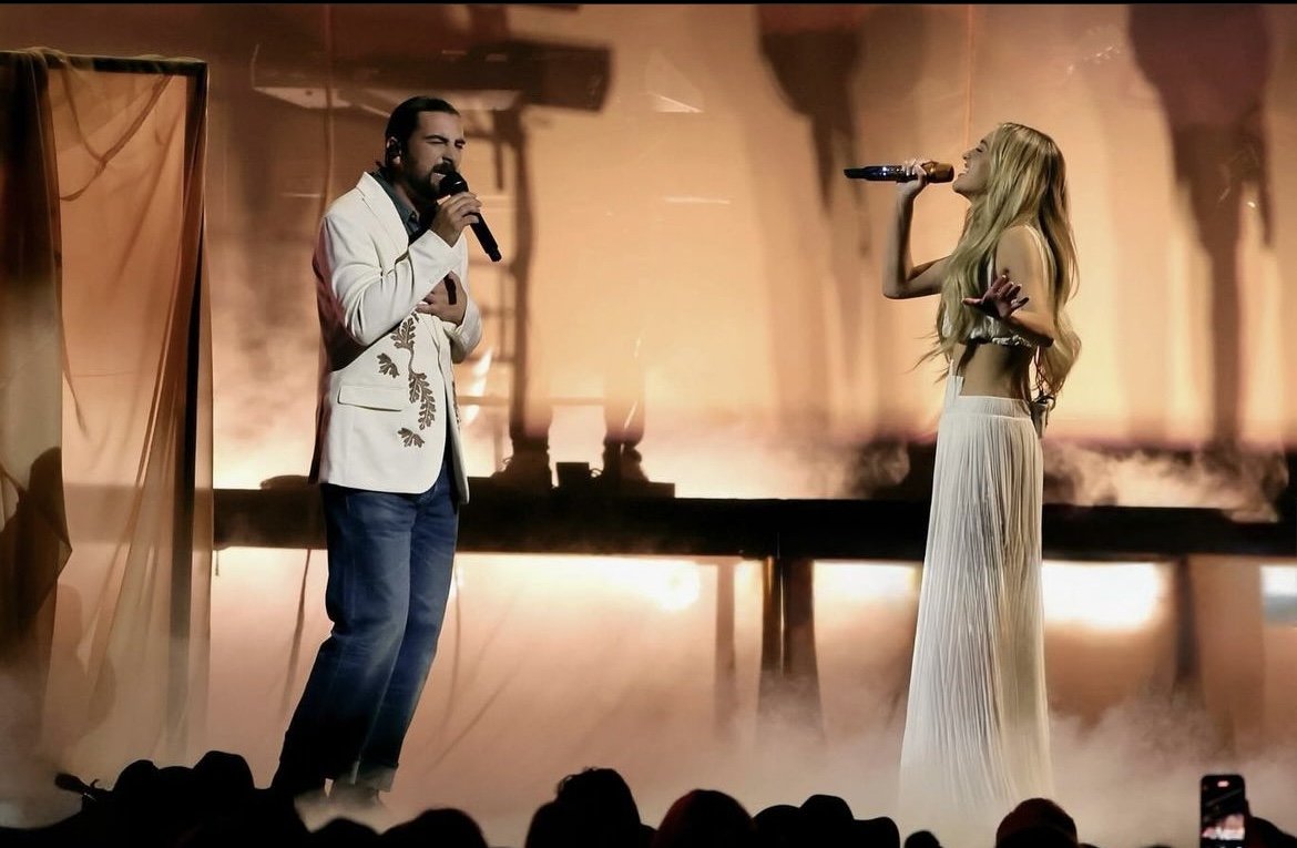 A man and woman singing on stage with microphones, surrounded by stage fog and warm lighting.