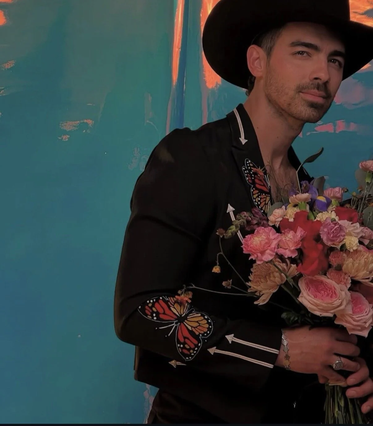 A man wearing a black cowboy hat and black shirt with floral embroidery and butterfly details, holding a colorful bouquet of flowers, standing in front of a teal wall.