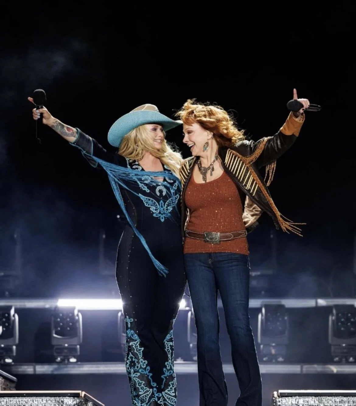 Two women are on stage, celebrating with their arms around each other, smiling, and holding microphones. One woman is wearing a wide-brimmed hat and decorative clothing, and the other is wearing a leather jacket with fringes and a red top.