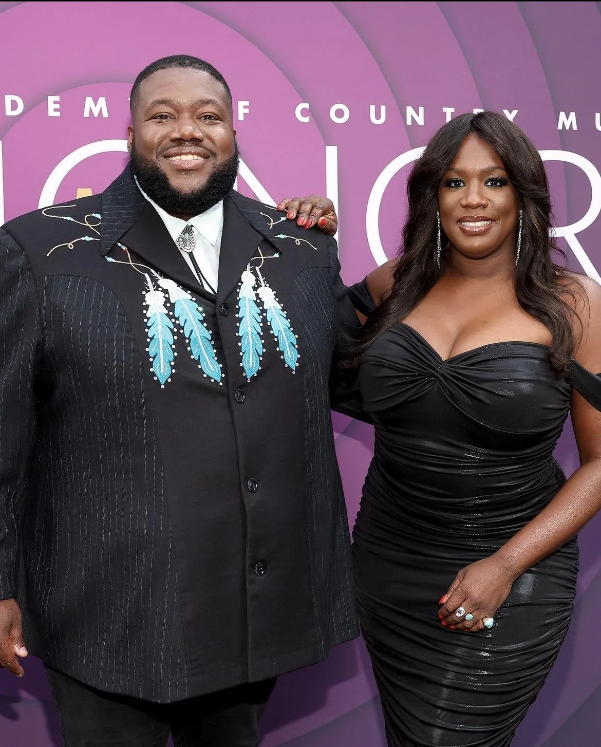 A man and a woman pose together in front of a purple backdrop at an event. The man is wearing a black suit with blue feather designs on the front, and the woman is dressed in a black, off-the-shoulder, ruched gown with long dark hair and jewelry.