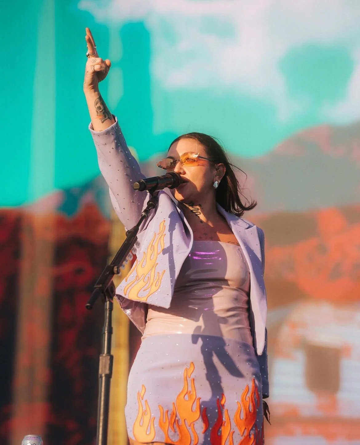 A woman performing on stage with a microphone, wearing sunglasses and a stylish outfit with flame designs, as she raises her hand during a concert with a desert landscape in the background.