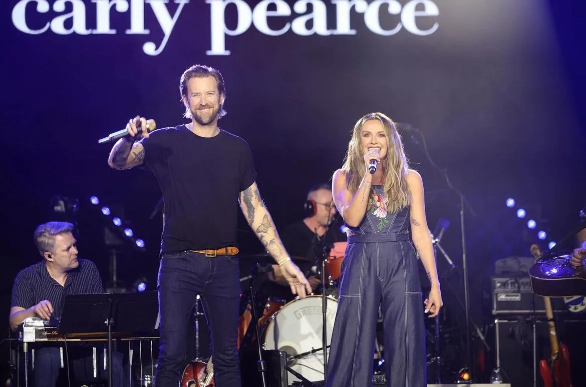 A man with tattoos and a woman singing into a microphone on stage during a concert with a backdrop that says "carry pearce". The man is smiling and holding a microphone in one hand, while the woman is smiling and holding a microphone in her right han