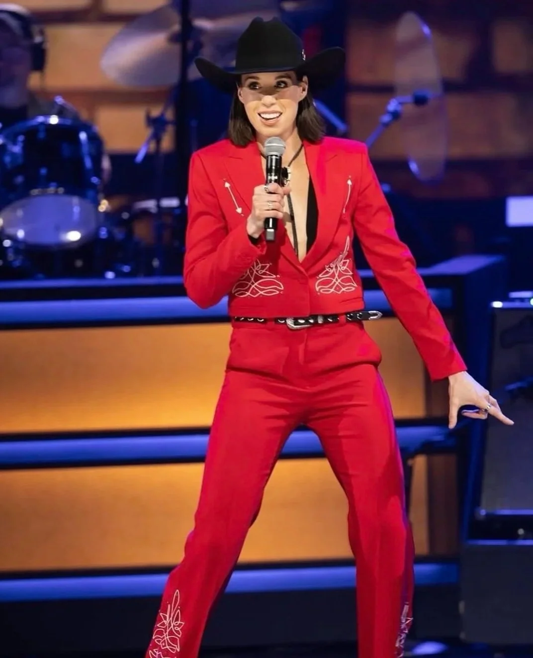A woman in a red suit with embroidery, wearing a black cowboy hat, holds a microphone and appears to be performing or speaking on stage, with drum set in the background.