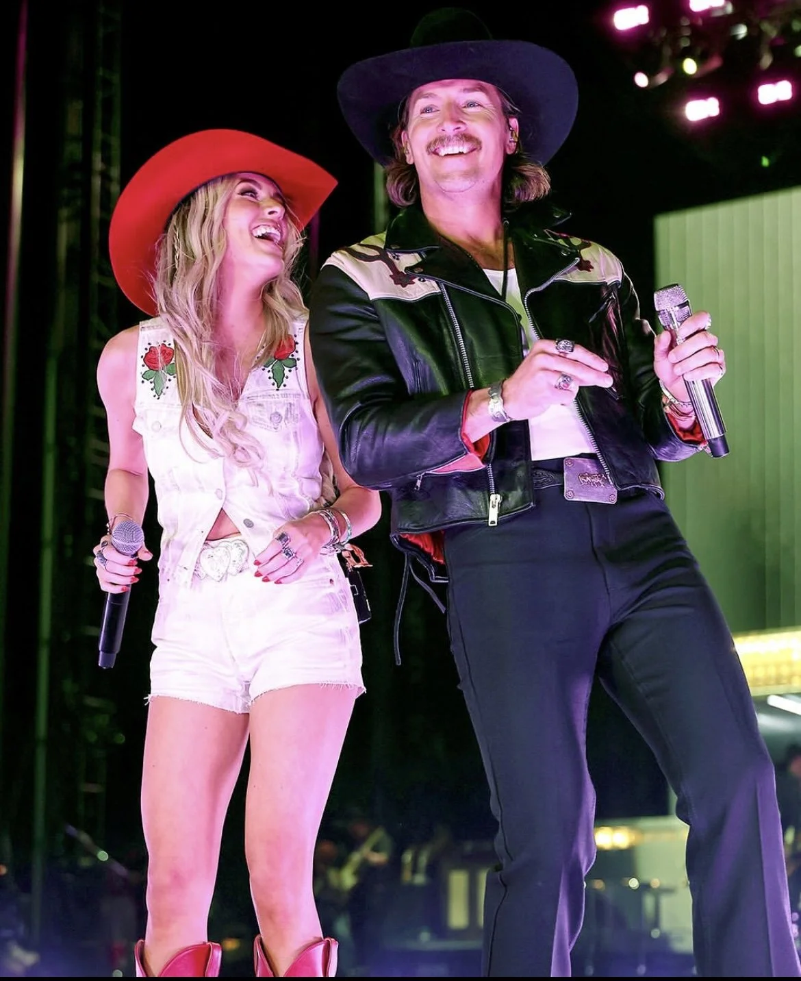 Two performers on stage, a smiling woman with long blonde hair in a white sleeveless top and shorts, and a man with long hair and a mustache wearing a black hat and leather jacket, both holding microphones.