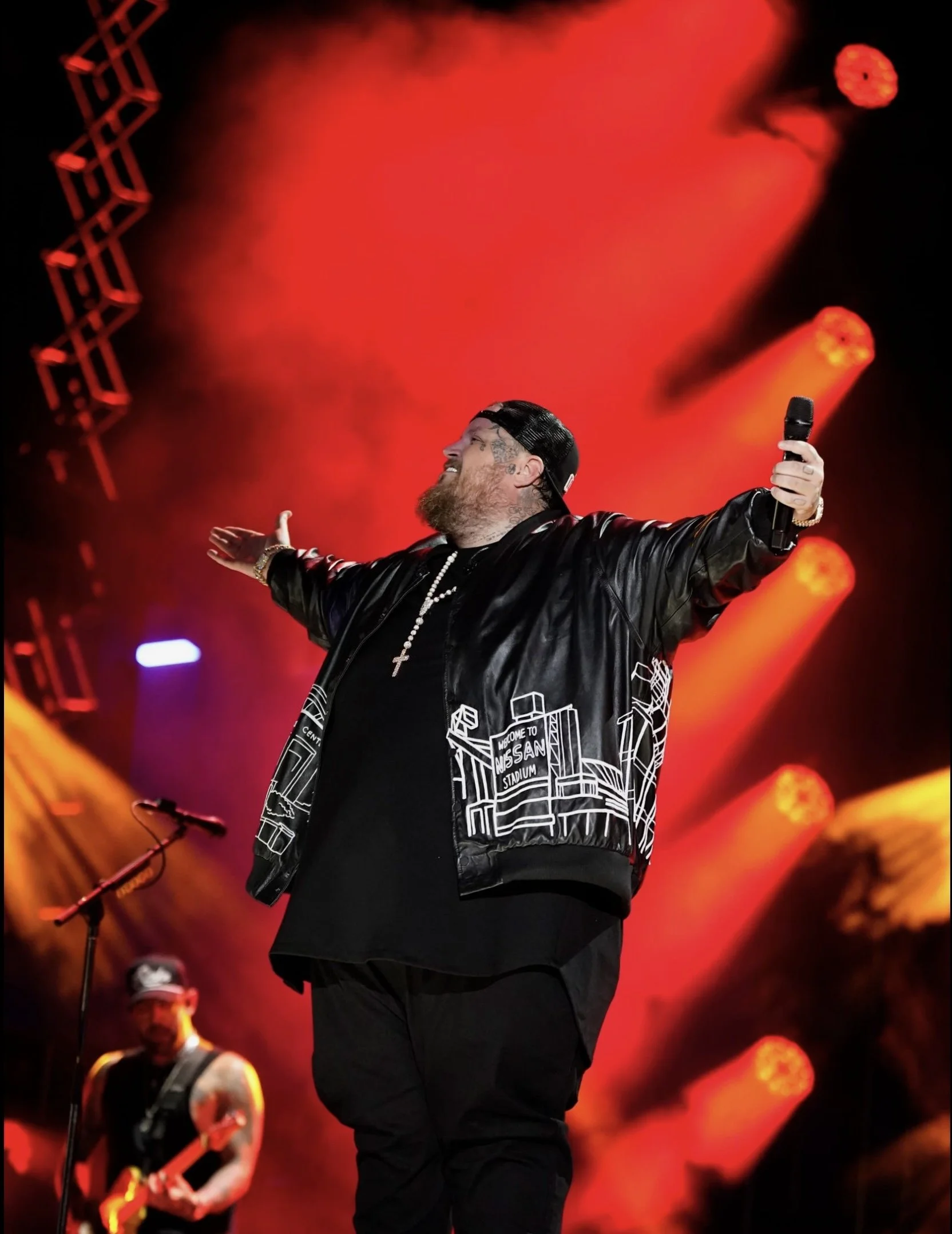 A man with a beard and tattoos on his face wearing a black cap, black jacket with a graphic design, and a white cross necklace, standing on stage with arms open and holding a microphone, with a red stage background and orange stage lights.
