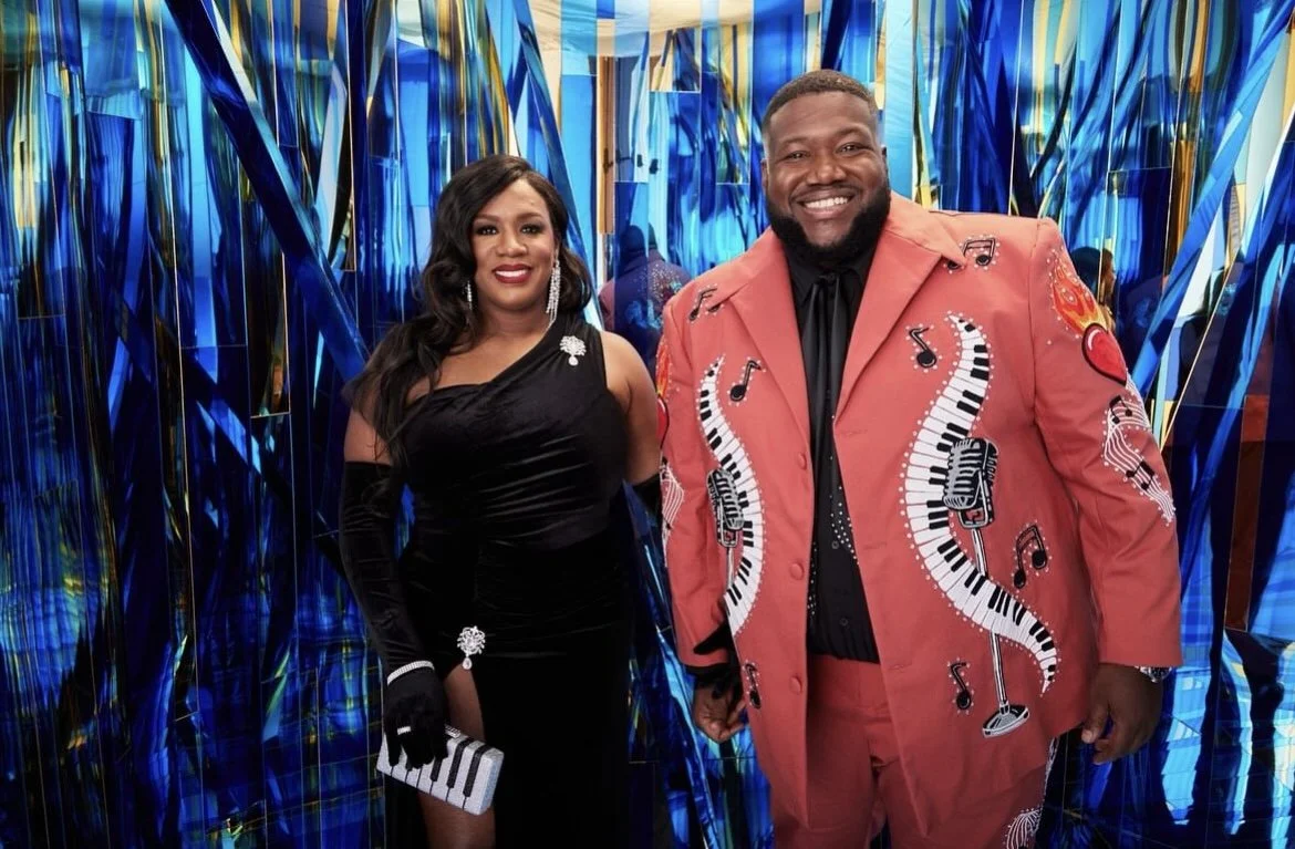 A woman and a man dressed in formal attire, standing in front of a blue abstract background. The woman wears a black dress with accessories, and the man wears a brightly colored jacket with musical note and piano key designs.