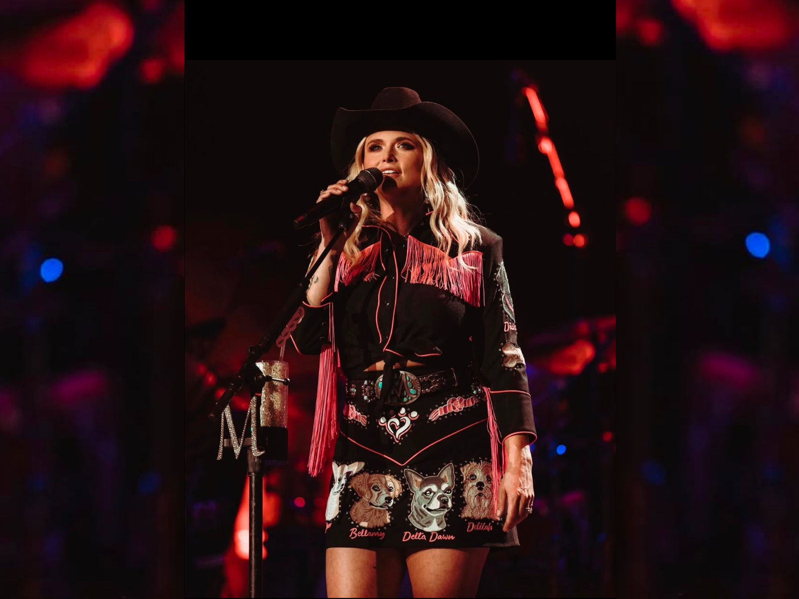 A woman dressed in a black and red Western-style outfit with fringe and animal patches, wearing a black cowboy hat, standing on stage holding a microphone, with a blurred colorful background.