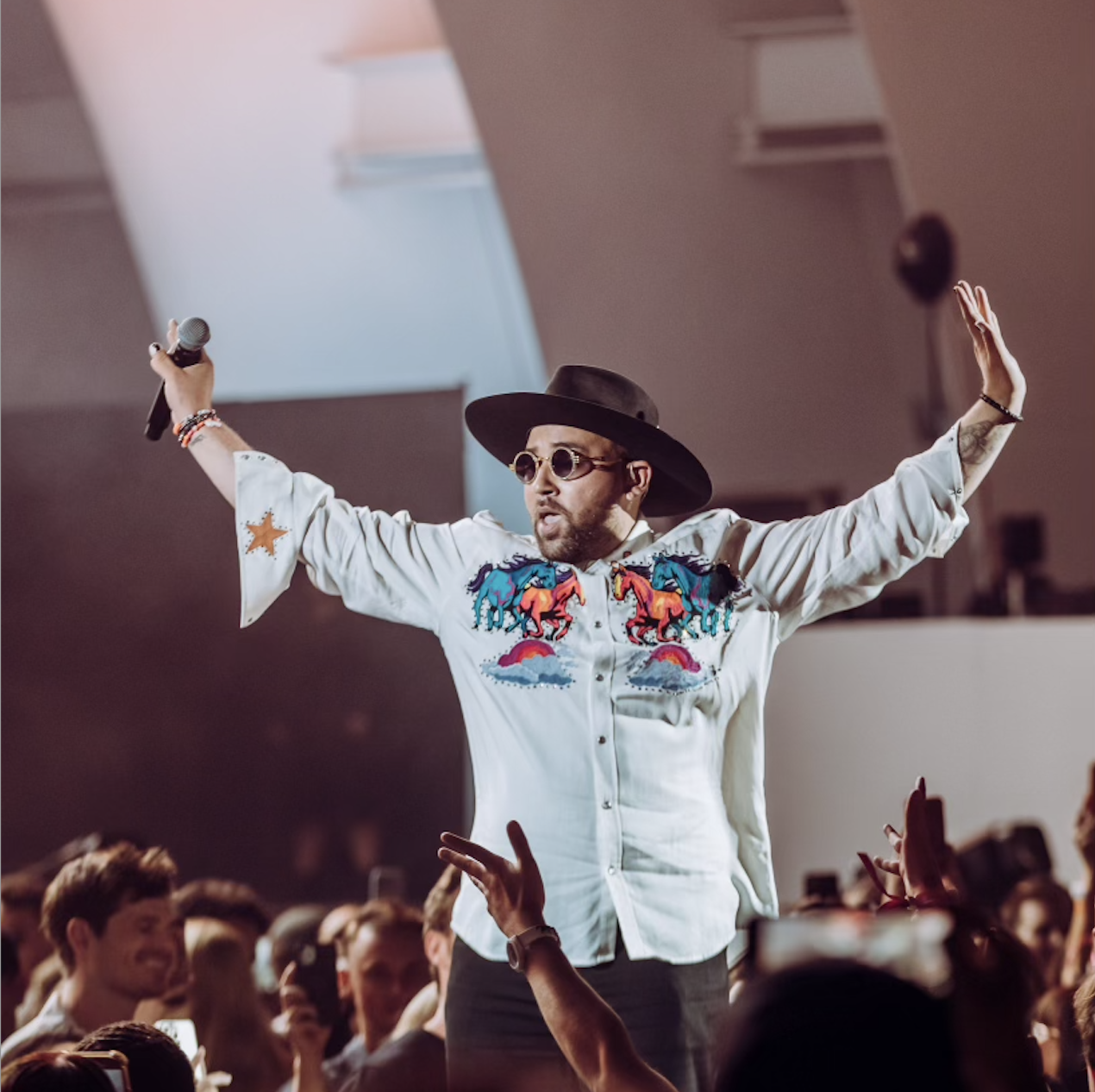 A man in a wide-brimmed black hat, sunglasses, and a white embroidered shirt with colorful horse designs stands with arms outstretched while holding a microphone, surrounded by a cheering crowd.