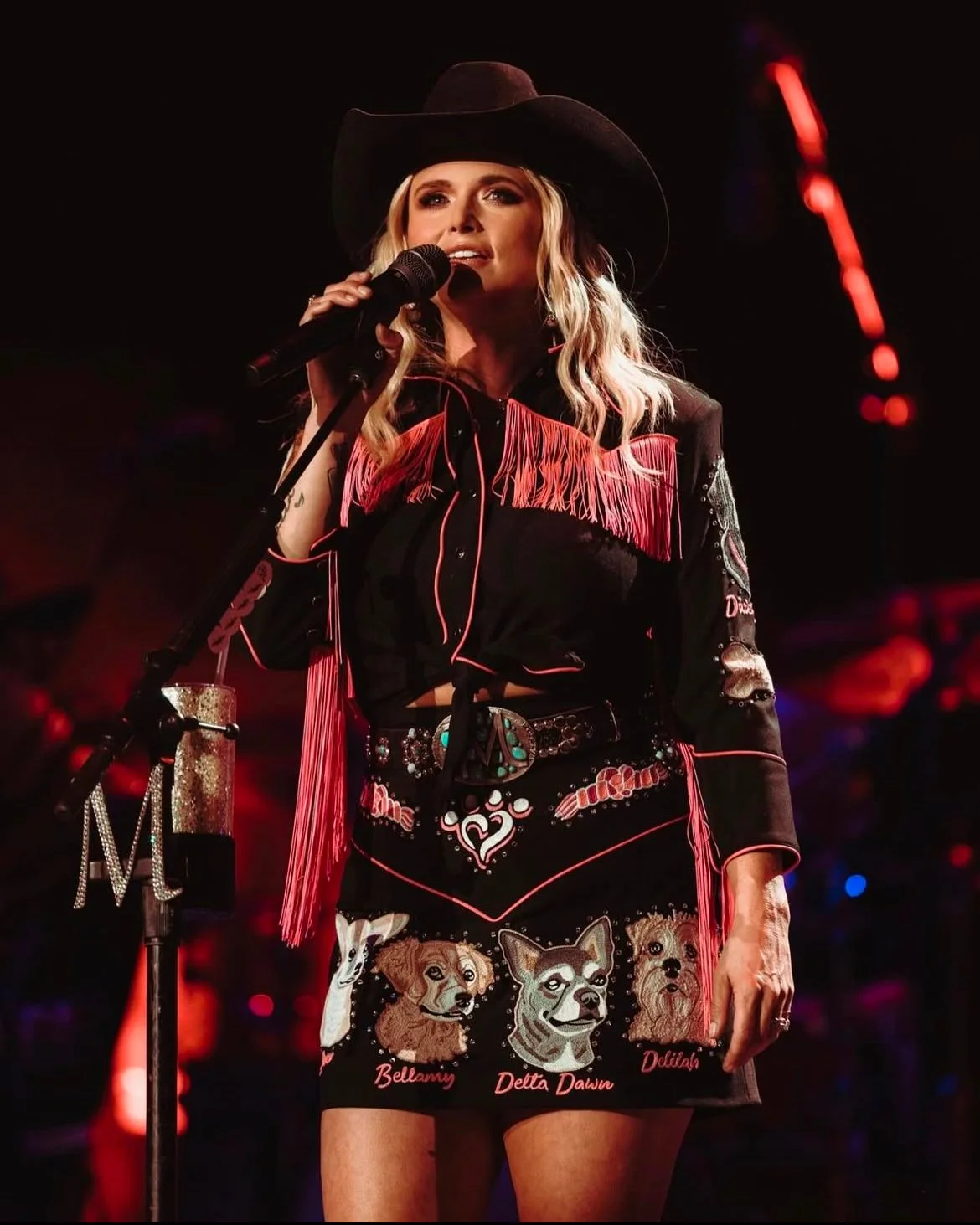 A woman dressed in a cowgirl outfit singing into a microphone on stage with red and purple lighting.