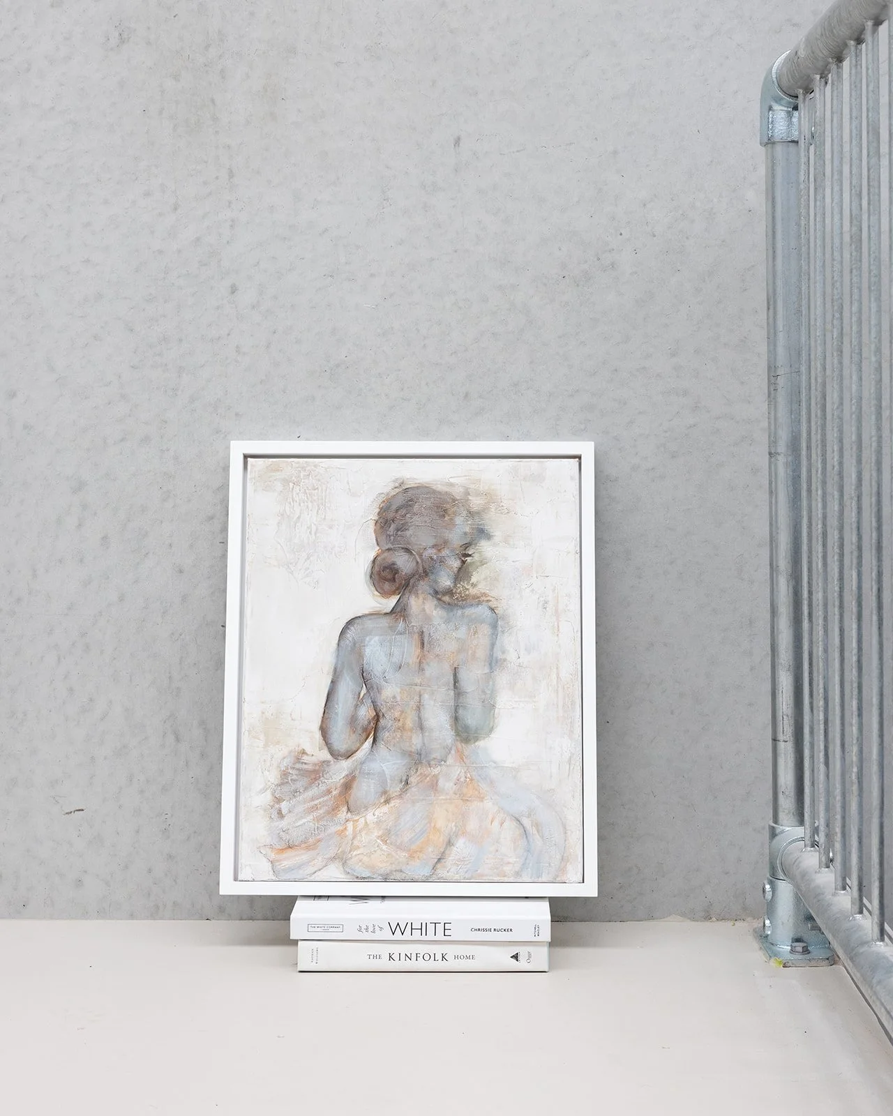 A framed painting of a young girl with a bun hairstyle sitting with her back turned, in soft muted colors. The frame rests on a stack of three art and design books, against a plain gray wall with an industrial metal railing on the right.
