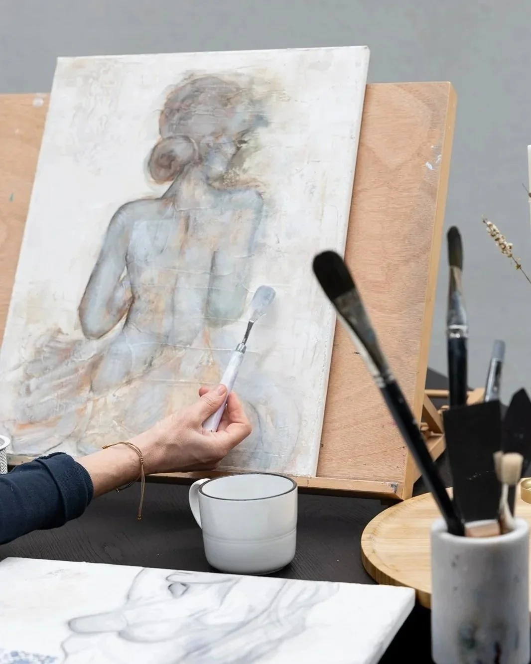 An artist is painting a portrait of a woman on a canvas. The artist holds a paintbrush near the canvas, which is propped on an easel. Various painting tools are visible, including brushes in a white container and a wooden board.