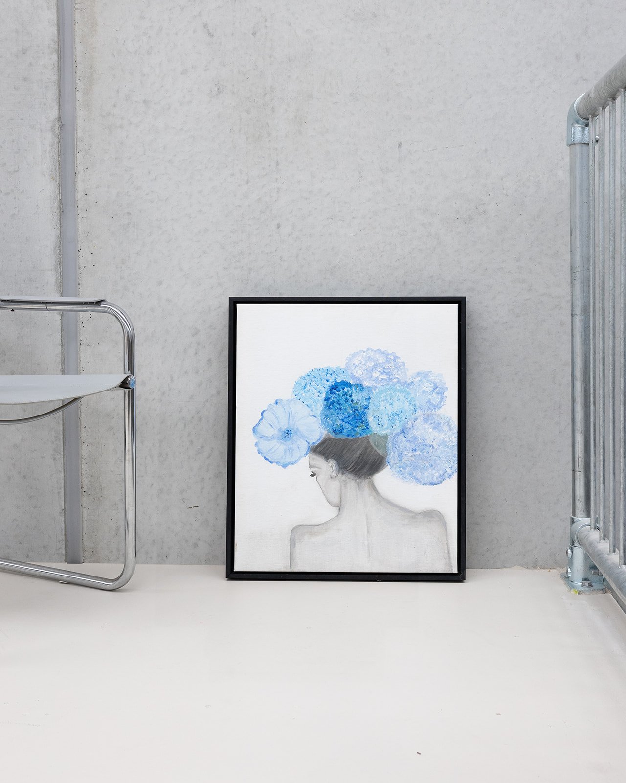 A framed artwork of a woman with her head turned to the side, adorned with a large bouquet of blue hydrangeas on her head, resting against a gray wall on the floor.