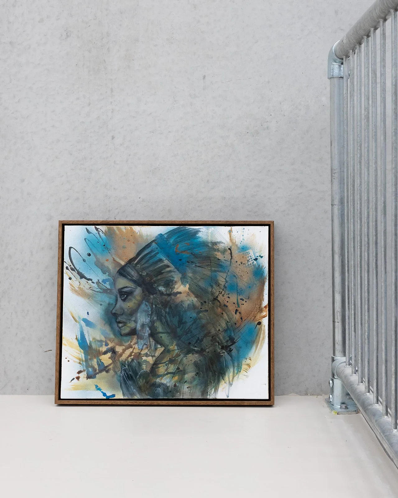 A framed abstract painting of a woman's profile with long hair, created with blue, brown, black, and white paint splashes, leaning against a textured wall near a metal railing.