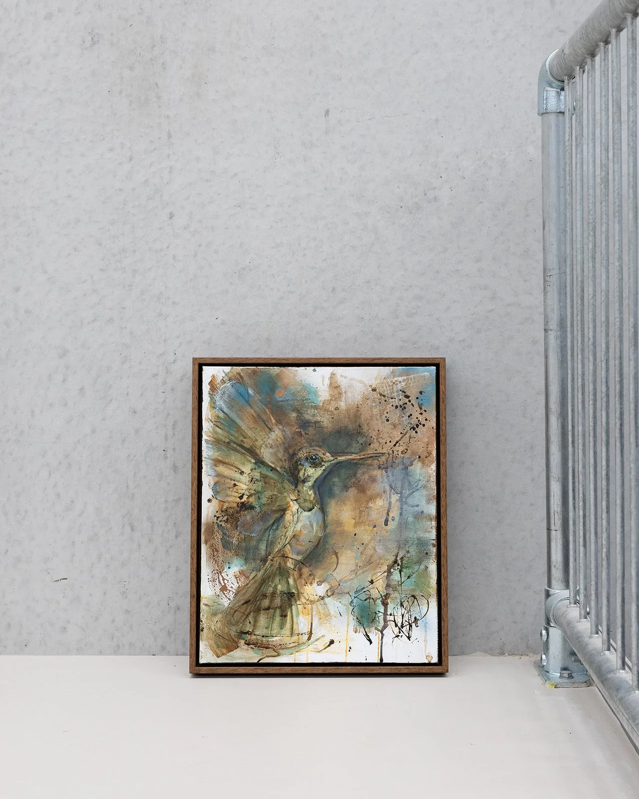Oil & Acrylic painting of a hummingbird with brown, ochre, teal, and metallic tones, leaning on a white surface against a textured light grey wall beside a metal railing.