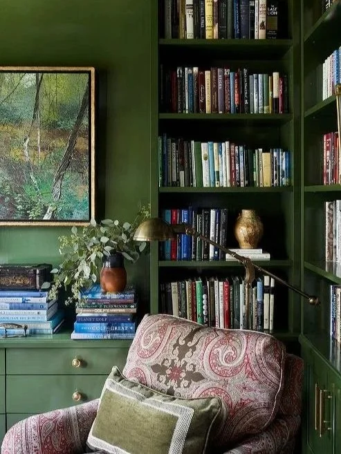 A cozy reading corner with green built-in bookshelves filled with books, a large framed landscape painting, a green armchair with a patterned cover and a pillow, a brass adjustable reading lamp, and a side cabinet with a stack of books and decorative vases.