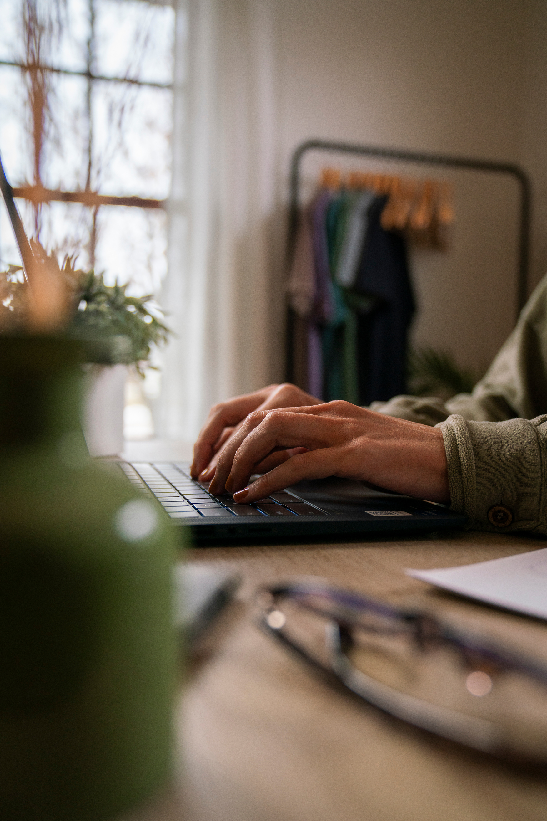 Close-up of a person's hands typing on a laptop keyboard on a wooden desk, with a blurred background including a window, clothing rack, and plant.