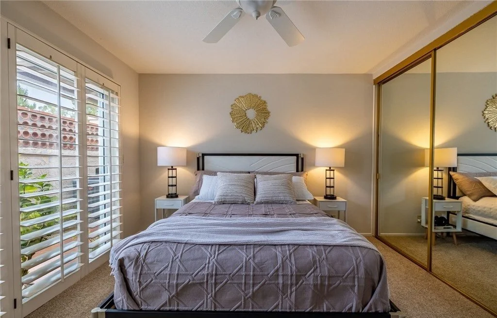 Paired lamps and sunburst artwork center the bed on its wall and draw the eye away from the mirrored closet doors, which are the room's most dated feature. Layered soft-lavender bedding and the light through the plantation shutters keep a smaller bed