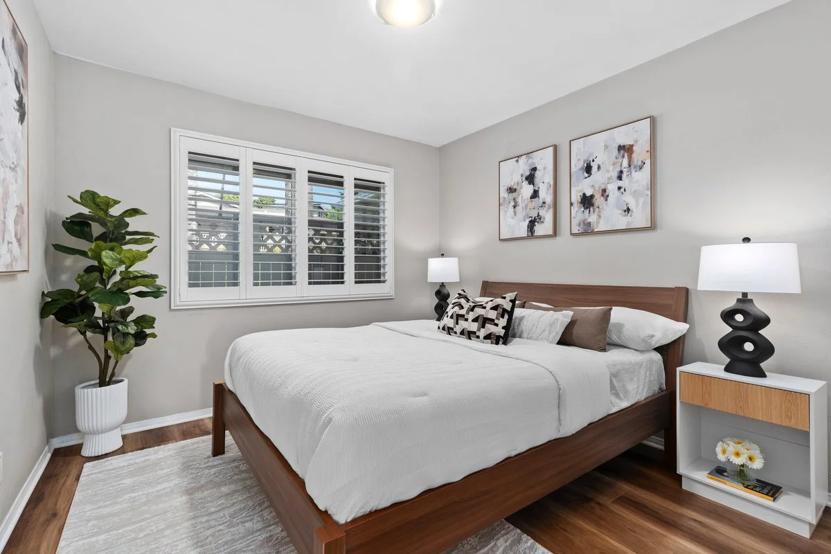 A walnut frame and a low case-goods nightstand pick up the mid-century lines the 1964 house already had. Two prints hung close above the headboard bring the eye down from a tall wall, and a single black lamp gives the pale bedding something to push a