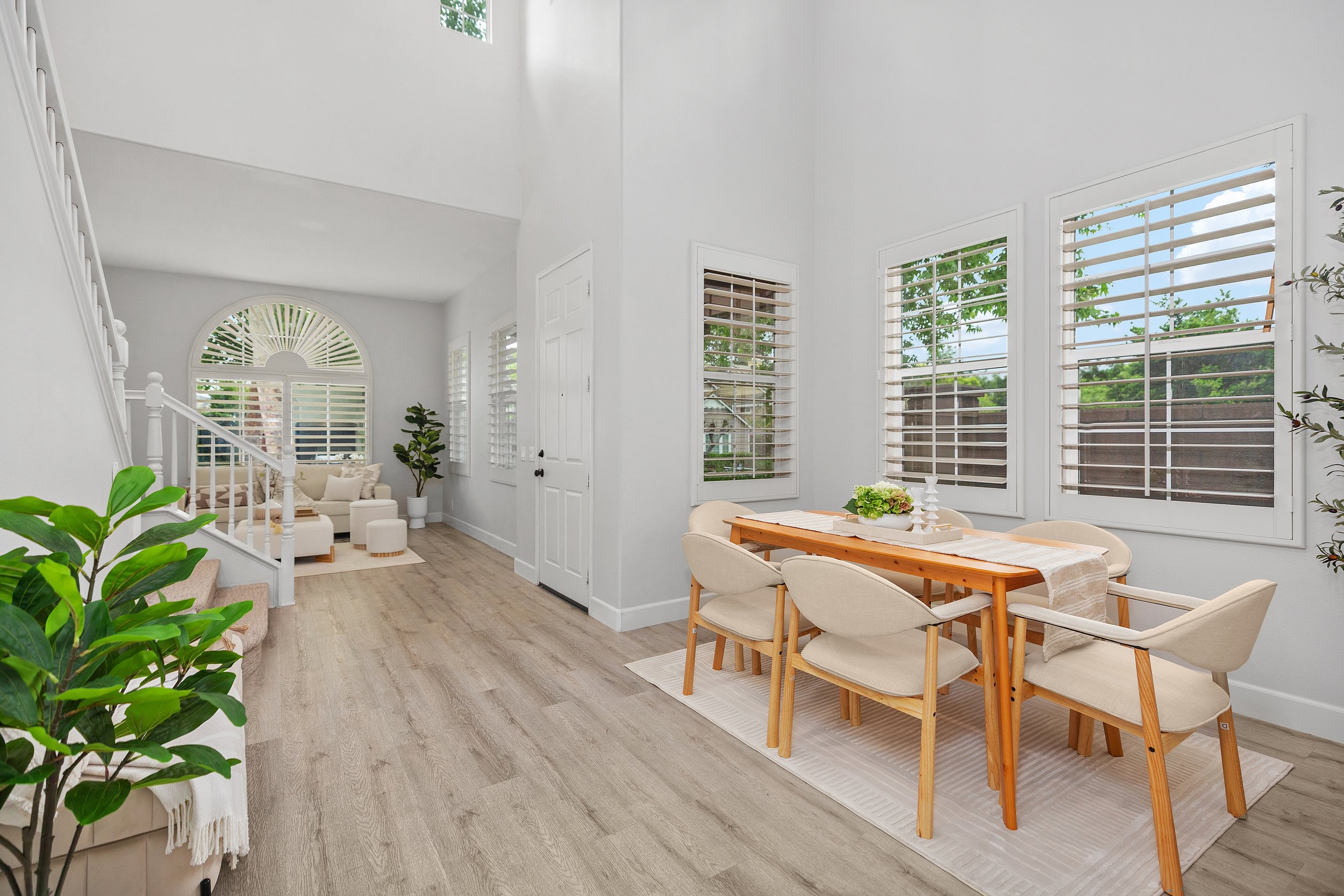 Bright dining area with wooden table and six beige chairs, large windows with blinds, white walls, view of greenery outside.