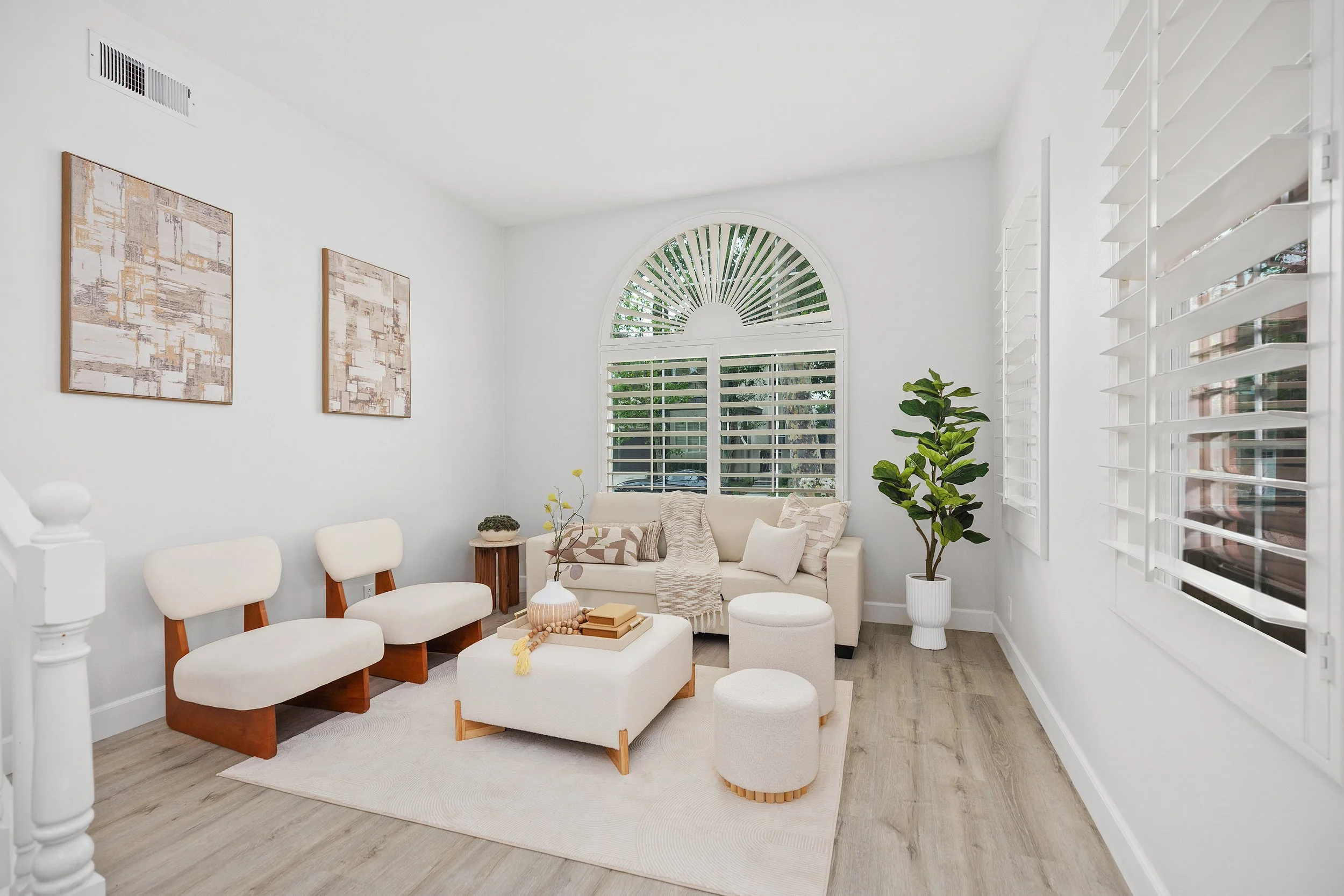 Bright living room with white walls, large arched window with white shutters, beige sofa with pillows, three white ottomans, wooden side table, two cream chairs, potted plant, wall art, and light wood flooring.