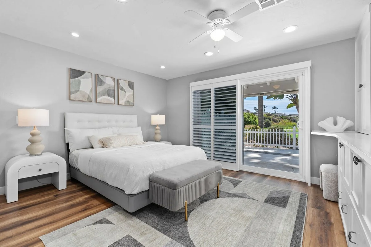 An upholstered bed centred between the nightstands, with a bench at the foot so the room doesn't end abruptly at the slider. Lamps instead of overhead light, and nothing tall on the window side, so the shuttered doors stay the brightest thing in the 