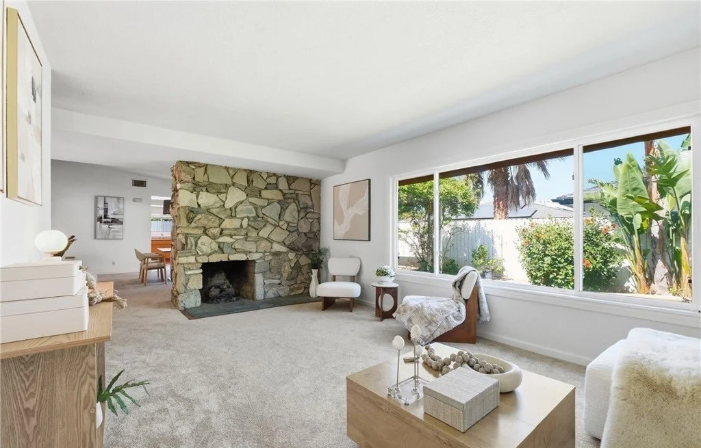 The original stone fireplace anchors this room, and the staging keeps everything around it low and light so nothing competes with it. An oak coffee table, textured seating, and a console in the foreground build depth, while the window wall holds the 