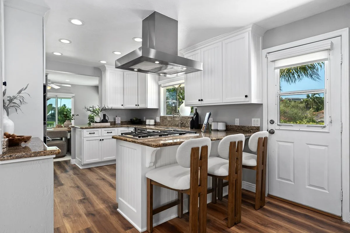 Three stools at the peninsula are the only furniture the kitchen needed. The granite is a busy speckle, so the accessories stay white, matte and few, and the cabinetry and the windows carry the room instead of competing with the counters.