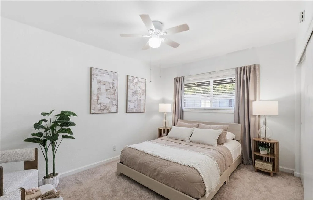Layered taupe bedding, paired lamps, and a matched set of framed abstracts give this secondary bedroom a symmetrical, restful setup. Full-length drapery softens the window line and adds height, while a corner plant and one of a pair of accent chairs 
