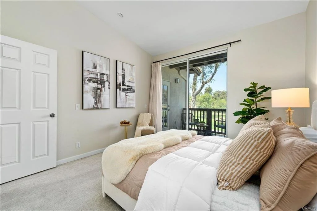 Layered neutral bedding, warm blush accents, and symmetrical styling create a serene bedroom designed to feel both luxurious and approachable. The adjacent balcony and natural light enhance the sense of a private Southern California retreat.