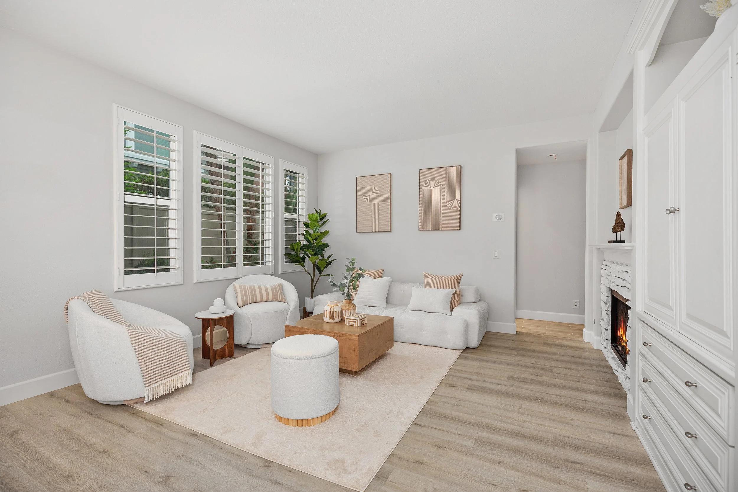 Bright living room with white walls, a white sofa with pillows, armchair, and a wooden coffee table, large windows with shutters, a fireplace, decorative art, and a potted plant.