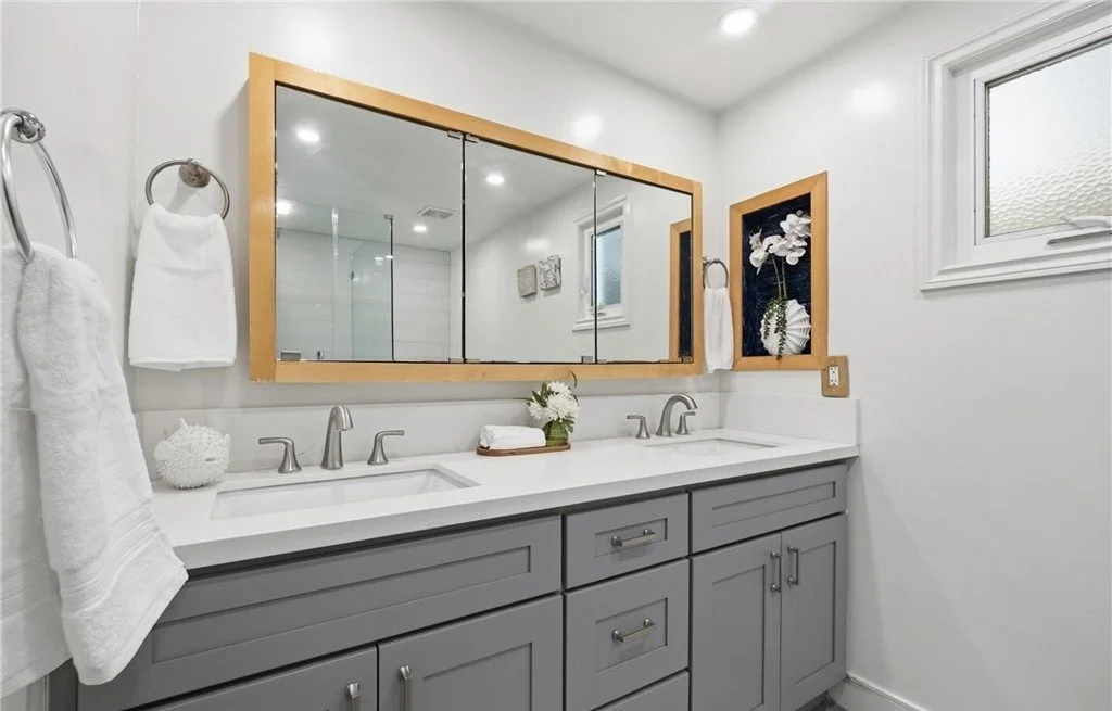 Styling here is intentionally minimal - rolled white towels, a small floral accent, and one botanical print are enough to finish the double vanity without hiding the counter space buyers came to see. The oversized wood-framed mirror carries warmth in