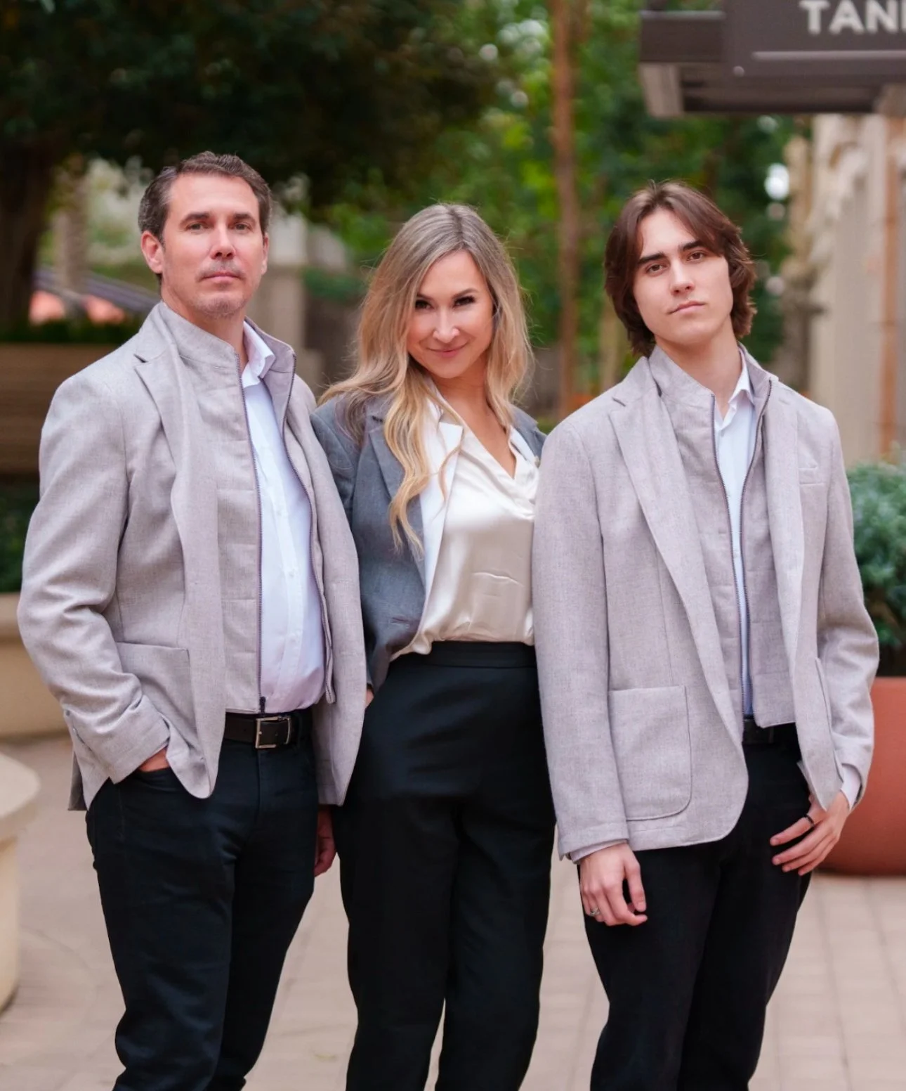 Three people standing outdoors, wearing light-colored blazers, with a greenery background.