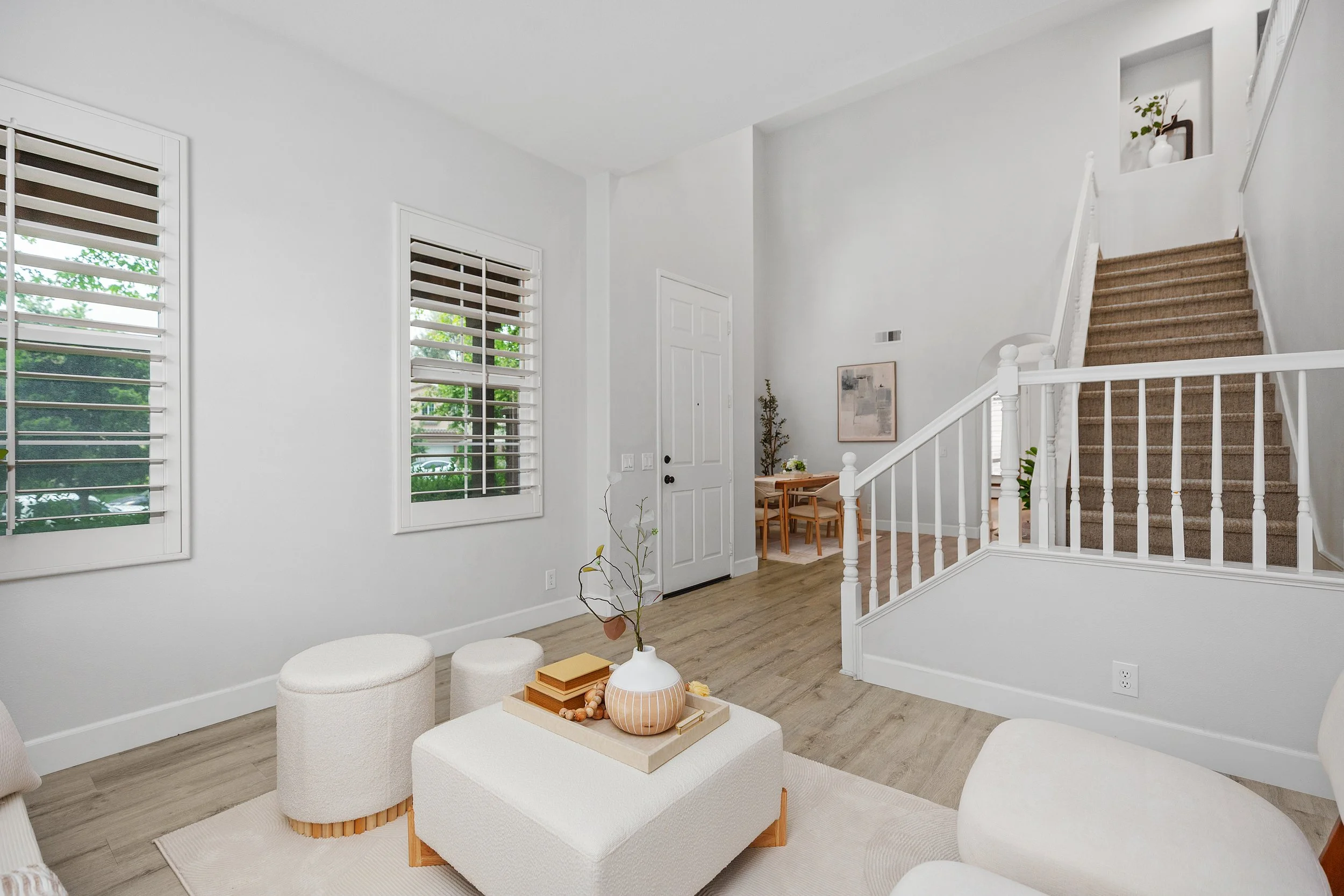 Bright living room with white walls, two large windows with shutters, a staircase with a brown carpet, a white and beige chair, and a small green plant on the wall.
