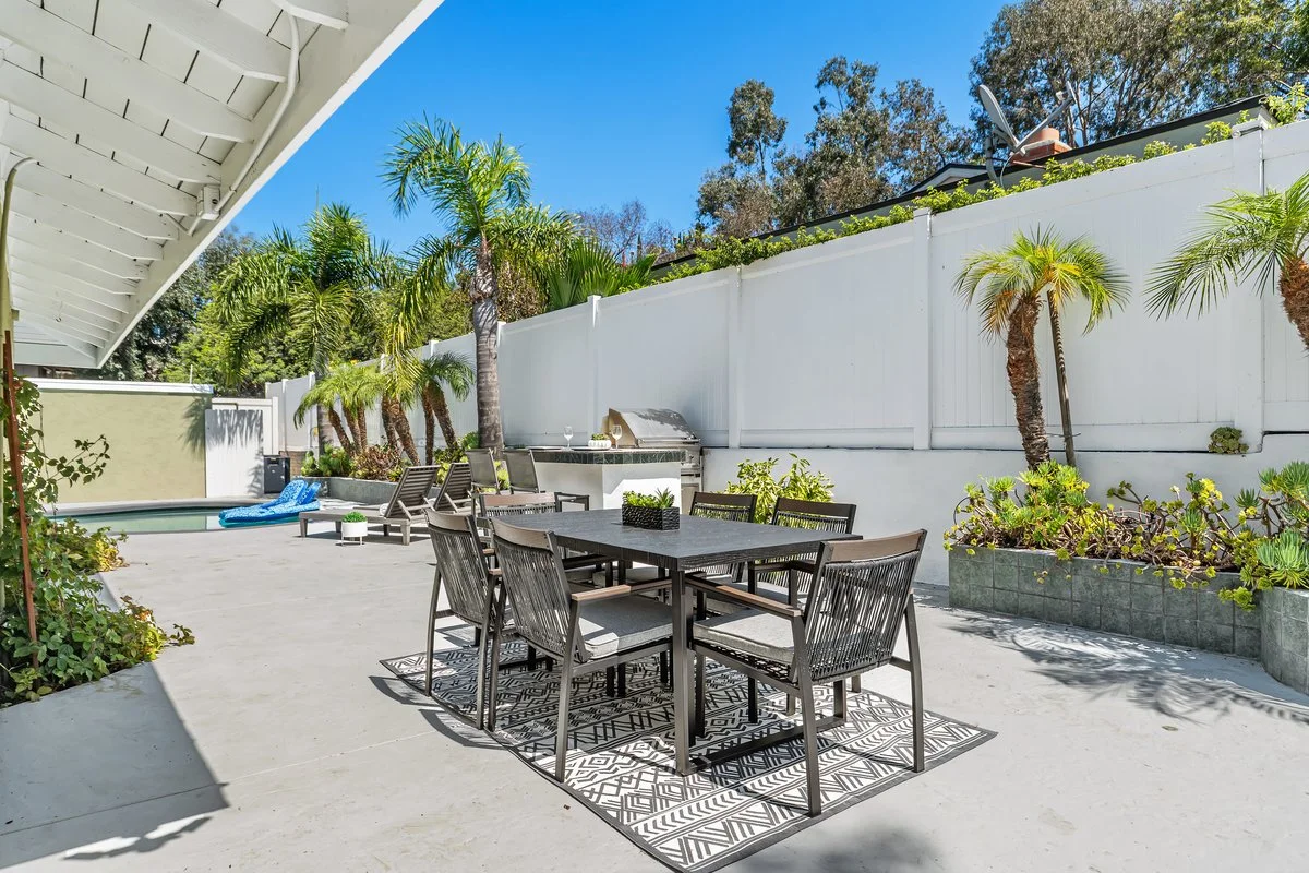 At the pool end of the same run, a six-seat dining set on an outdoor rug with two loungers turned toward the water. The rug is doing the work here: on an unbroken expanse of concrete it is what tells you where the dining room is.