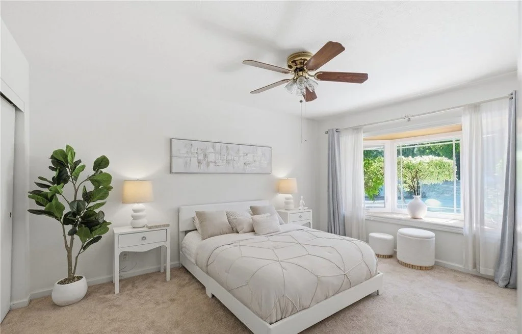 A crisp white bed frame and quilted cream bedding keep the primary bedroom feeling light, with matching nightstands and lamps establishing symmetry on a modest wall. Sheer curtains filter the daylight coming through the front window, and a tall corne