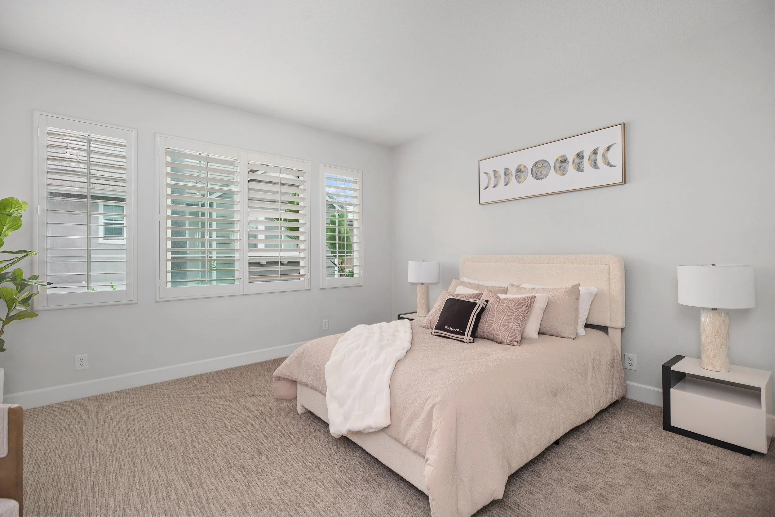 A bedroom with a beige upholstered bed, white bedding, and multiple pillows, flanked by white nightstands with lamps, white walls, a framed moon phase art piece, and a large window with white plantation shutters.