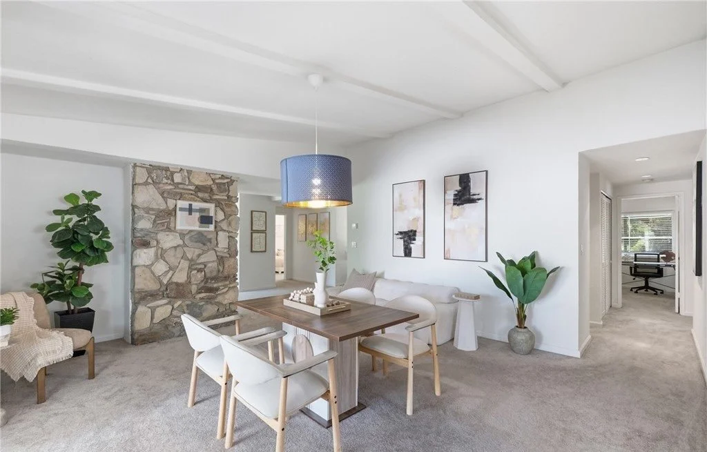 A warm wood dining table, upholstered chairs, and a low couch tucked in behind them keep the seating soft and informal against the home's original stone wall, while a single blue pendant gives the neutral palette one deliberate point of color. Greene