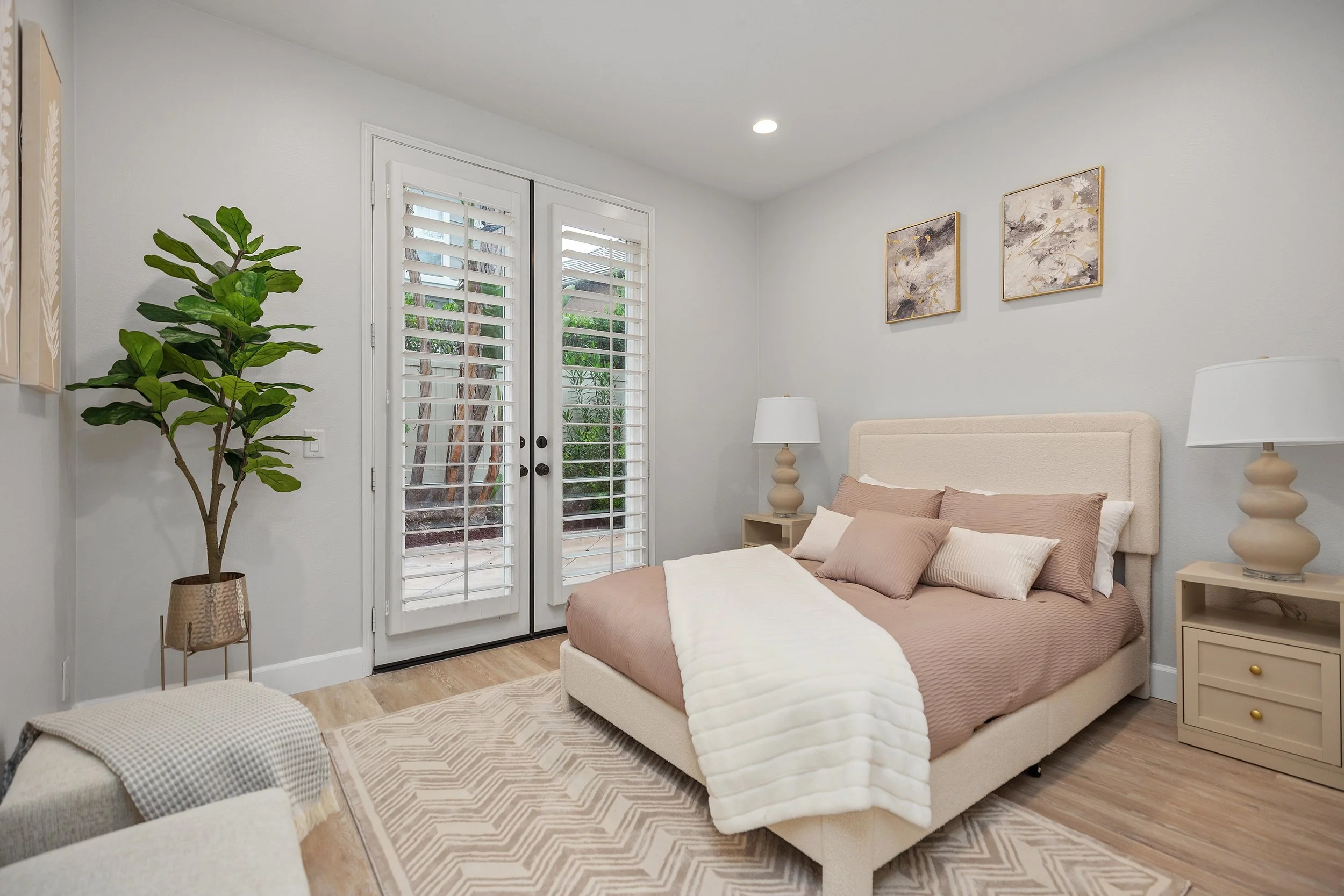 Bedroom with large bed, beige headboard, white and beige bedding, two nightstands with lamps, water and plant outside through glass doors, light-colored walls, artwork above bed, wooden flooring, stress with pattern, large potted plant in corner.
