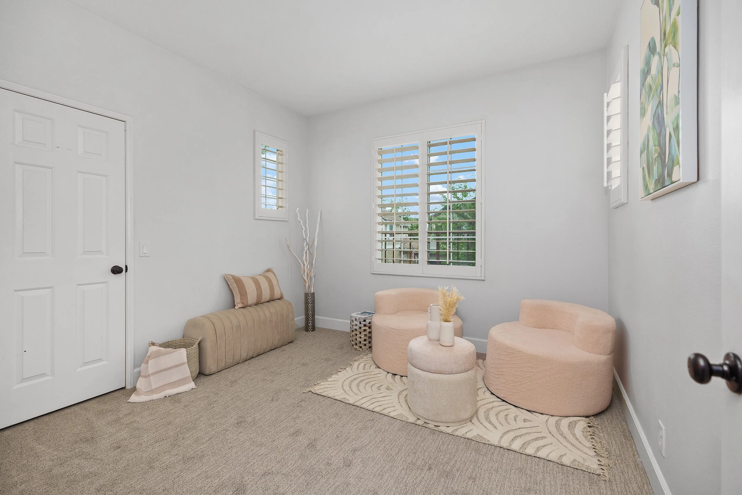 A small, minimalist living room with white walls, carpeted floor, and large windows with white shutters, decorated with pink and beige furniture and abstract art.