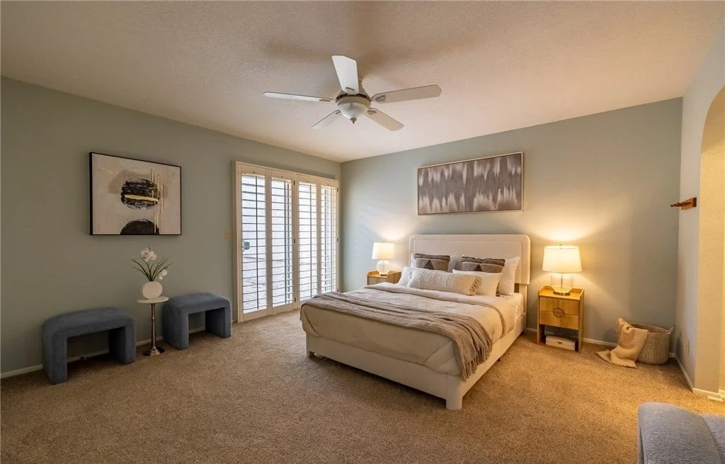 Sage walls and a low upholstered bed keep the primary bedroom calm, with matched lamps and a single wide canvas above the headboard establishing symmetry on the main wall. A pair of dark ottomans by the window fills the corner and gives the room a se