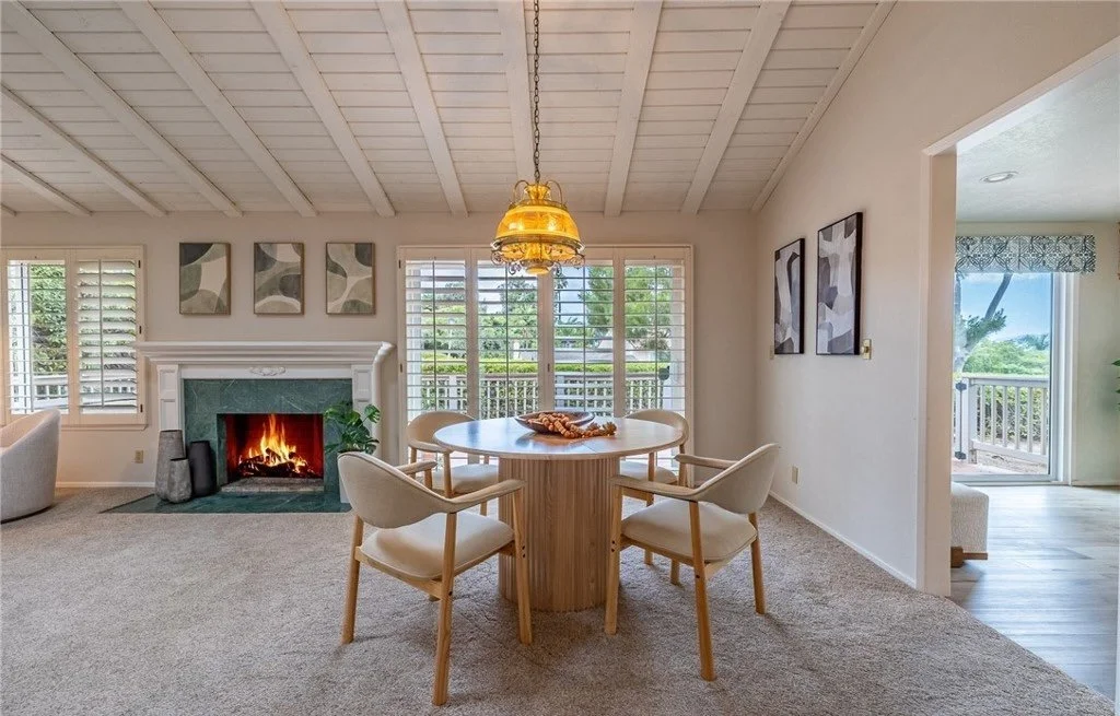 A round pedestal table seats four without blocking the traffic path between the fireplace and the balcony doors, and the brass pendant marks the dining zone in a room that has no walls to define it. Light wood chairs keep the setting from competing w