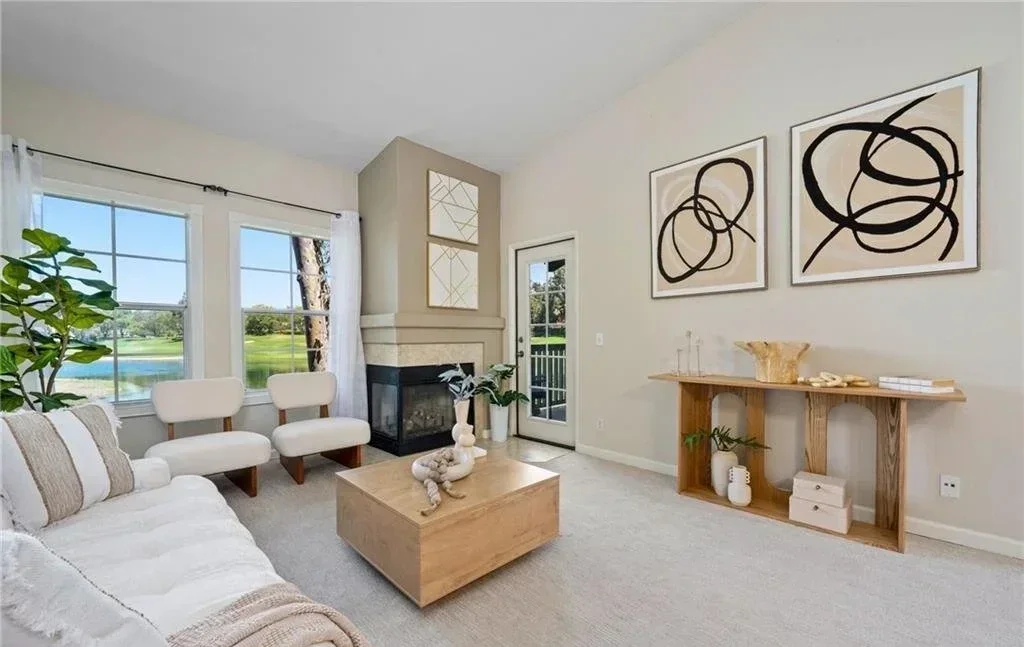 Paired accent chairs and a low oak coffee table pull the eye toward the fireplace, while a slim console behind the sofa adds height without closing the room off. Oversized abstract art and layered neutrals keep the vaulted living space calm and brigh