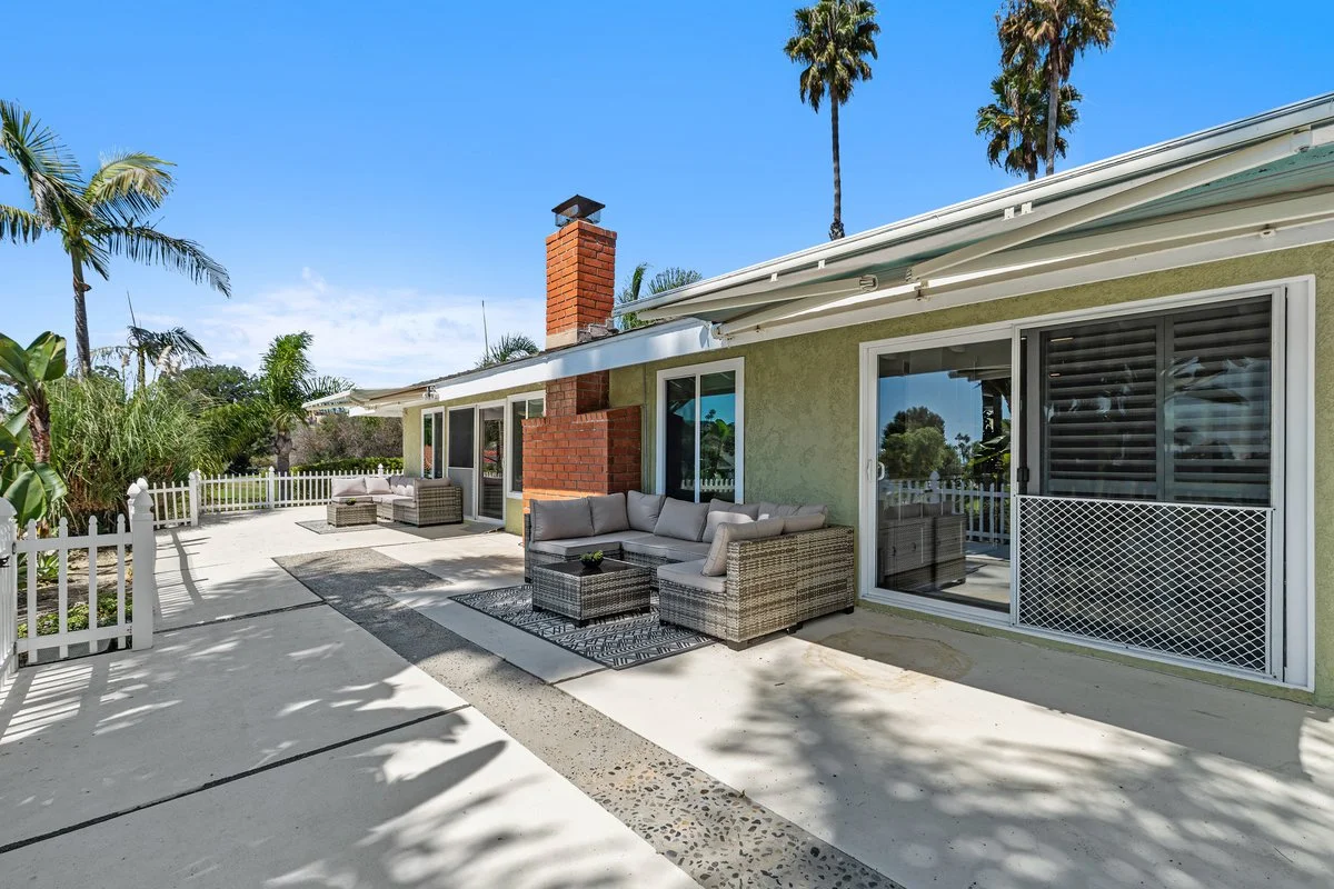 The patio ran the full length of the house as bare concrete. Two rattan groupings, one on each side of the brick chimney, break it into a pair of outdoor rooms instead of one undefined slab. Each sits on its own rug, so the furniture reads as placed 