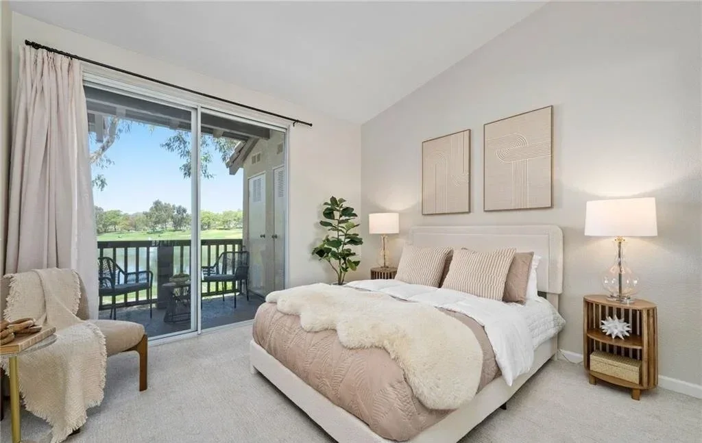 Sliding doors open the bedroom onto a private balcony overlooking the greenbelt and water, with the outdoor seating staged to read as a second living space rather than leftover square footage. Inside, a corner reading chair and slim rattan nightstand
