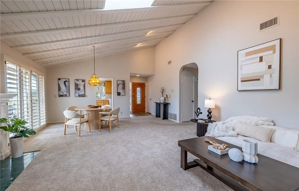 The seating is pulled to one end and kept low so the full length of the great room reads in a single look, from the fireplace wall through the dining area to the front door. A long, low table in front of the sofa anchors the arrangement without inter