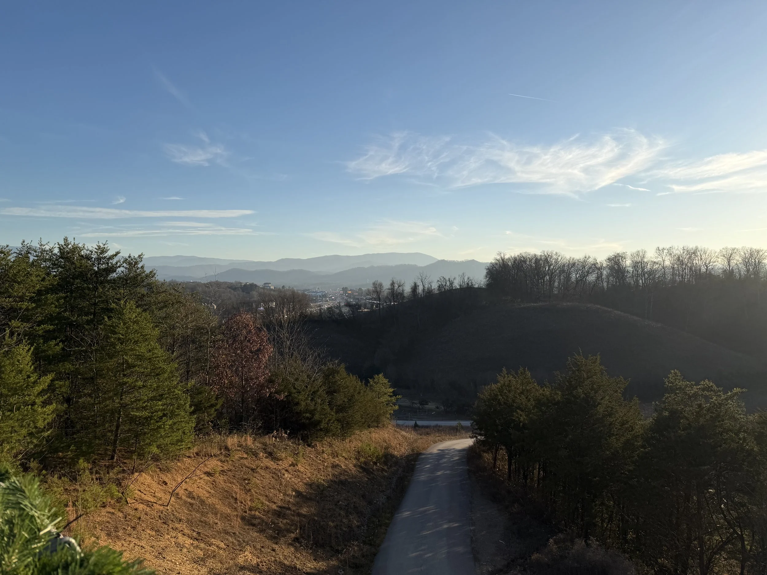 A winding dirt road through hilly terrain with trees, under a blue sky with wispy clouds and distant mountains in the background.