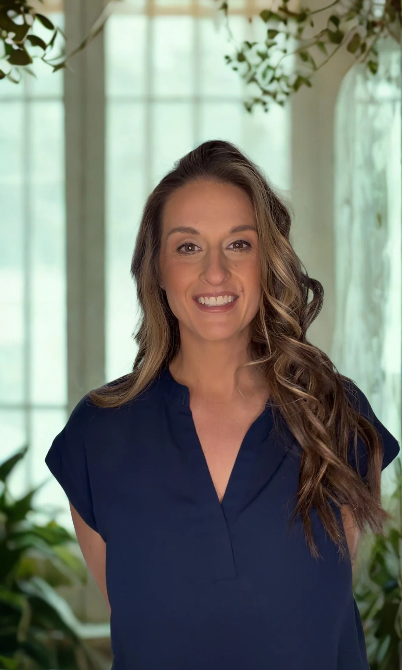 A woman with long wavy brown hair wearing a navy blue top, smiling indoors with sheer curtains and green plants in the background.