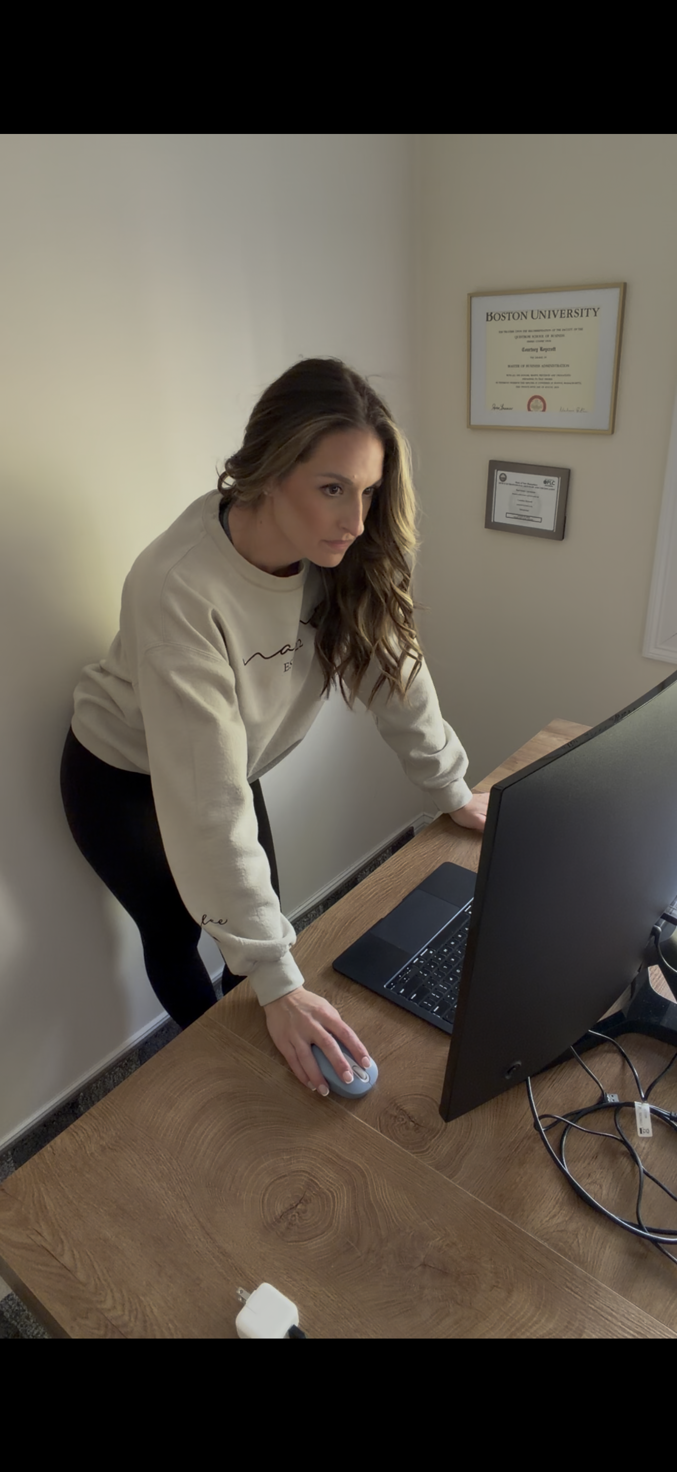 Woman with long wavy brown hair leaning over a wooden desk, using a computer mouse and looking at a monitor, with diplomas on the wall behind her.