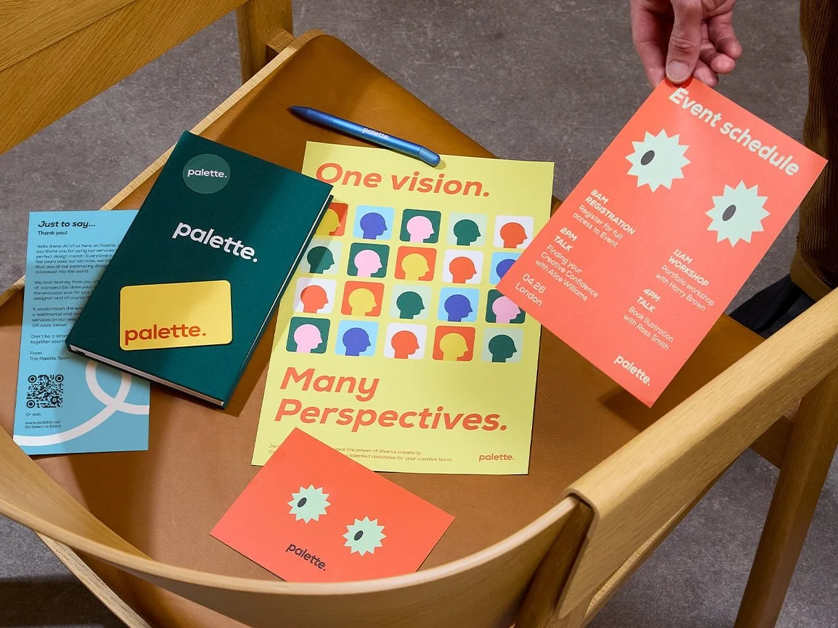 A wooden table with flyers, cards, and a pen promoting diversity and creativity. The flyer says 'One vision. Many Perspectives.' and features colorful head silhouettes. Two orange event flyers list scheduled activities, and a navy blue card with the word 'palette' appears on top of a blue informational card.